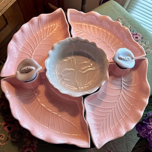 May include: A pink ceramic serving dish set. The set includes four leaf-shaped dishes surrounding a central bowl. Each leaf dish has a detailed vein pattern. Two small, white, peach-shaped accents are attached to the leaf dishes.