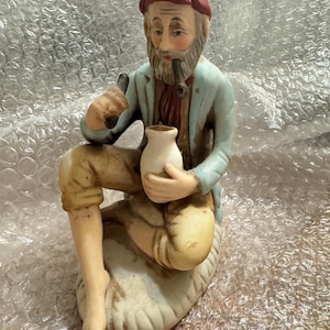 Könnte beinhalten: Keramikfigur eines alten Mannes mit Bart, roter Mütze, hellblauer Jacke und beigefarbener Hose. Er hält eine Pfeife und eine kleine weiße Vase. Die Figur sitzt auf einem cremefarbenen Sockel.