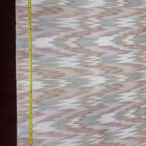 May include: A piece of fabric with a repeating zigzag pattern in shades of beige, white, light blue, and lavender. A yellow measuring tape is placed along the edge of the fabric, showing measurements in inches.