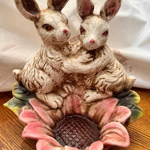 Puede incluir: Figura de cerámica con dos conejos abrazados, colocada sobre una flor grande. Los conejos tienen pelaje texturizado, narices rosas y ojos rojos. La flor tiene pétalos rosas, hojas verdes y un centro marrón oscuro. La figura tiene un esmalte craquelado.