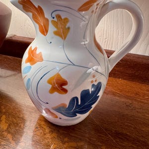 May include: White ceramic pitcher with a curved handle and spout. The pitcher is decorated with orange, yellow, and blue leaf designs and blue vine-like lines. The pitcher is a decorative item.