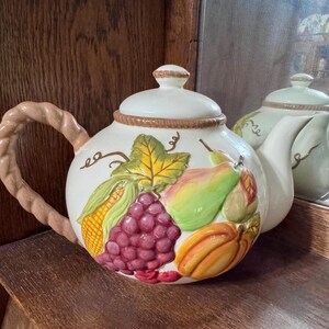 Op de afbeelding: Een witte keramische theepot met een fruitontwerp. De theepot heeft een gevlochten handvat en een deksel met een kleine knop. Het fruitontwerp omvat druiven, maïs, peren en een pompoen. Een tweede theepot is zichtbaar op de achtergrond.