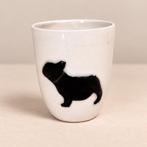 Puede incluir: Un vaso de cerámica blanca con una silueta negra brillante de un bulldog francés en la parte frontal. La taza tiene una forma cilíndrica ligeramente cónica con un borde limpio y un acabado blanco opaco y sólido en todo el exterior.