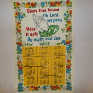 May include: A vintage 1985 kitchen towel with a floral border and a prayer for the home. The towel features a calendar for the year 1985 and the text "Bless this house Oh Lord, we pray, Make it safe By night and day."