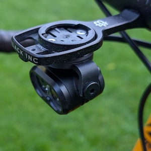 Könnte beinhalten: Schwarzer Fahrradscheinwerfer und Halterung am Lenker befestigt. Die Halterung ist mit "BLACK INC" beschriftet und hat ein kreisförmiges Design zur Aufnahme eines Fahrradcomputers. Das Licht hat zwei Linsen und ist unter der Halterung montiert.