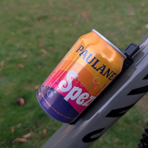 Könnte beinhalten: Ein graues Fahrrad mit einem schwarz-weißen Streifenmuster am Rahmen. Eine Dose Paulaner Spezi Orangensoda ist mit einer schwarzen und grauen Fahrradhalterung am Rahmen befestigt.