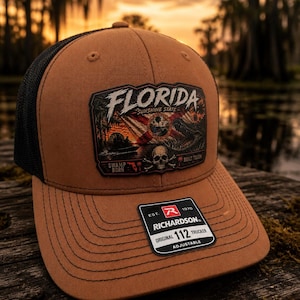 May include: A brown and black trucker hat with a patch that reads "FLORIDA SUNSHINE STATE". The patch features a skull, alligator, and swamp scene. The hat is adjustable and has the text "ORIGINAL 112 TRUCKER".