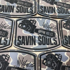 May include: A set of five black and white patches with a camouflage background. The patches feature a chainsaw with the text "RIPPIN HOLES SAVIN SOULS".