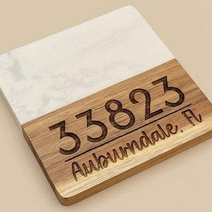 May include: Square coaster with a white marble-like top and a wooden bottom. The wooden section is engraved with the numbers "33823" and the word "Auburndale, FL".