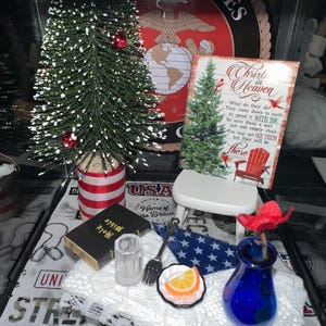 May include: A miniature Christmas display with a small green Christmas tree, red baubles, and a blue vase with a red flower. Includes a miniature Bible, a fork, and a plate with a slice of orange. The display has a patriotic theme.