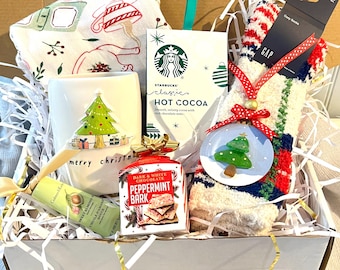 May include: A Christmas-themed gift box filled with a white mug featuring a Christmas tree design, Starbucks hot cocoa, peppermint bark, a pair of cozy socks, and a sweet almond hand cream. A festive holiday gift.