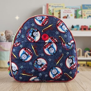 May include: A dark blue denim-style backpack featuring a repeating pattern of Thomas the Tank Engine in circular frames, interspersed with small light blue stars and the word Thomas in yellow script, finished with a single red button closure.