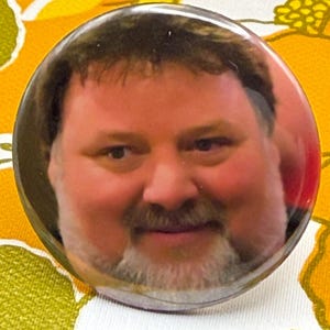 May include: A round button featuring a man's face with a beard and mustache. The button is set against a background of a floral pattern in shades of yellow, white, and green.
