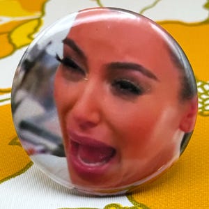 May include: A round button with a glossy finish featuring a close-up image of a person with a distressed expression. The button is set against a yellow and white floral patterned background.