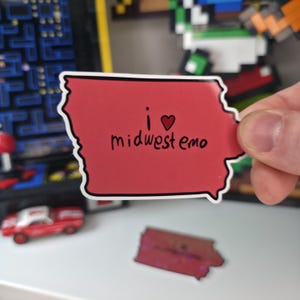 May include: A red sticker in the shape of Iowa with the words "i ❤️ midwest emo" in black script. The sticker has a white border. Other items are visible in the background, including a toy car and pixel art.