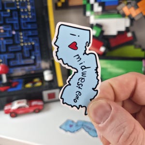 May include: A light blue sticker in the shape of the state of New Jersey with a red heart and the words "midwest emo" in a handwritten font. The sticker is held in a person's hand.