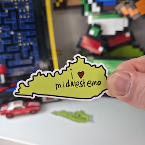 May include: A sticker in the shape of the state of Kentucky, colored yellow-green, with the words "i ❤️ midwest emo" in black and a red heart. The sticker is held by a hand.