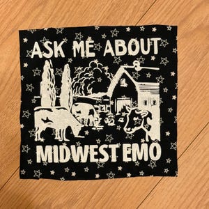 May include: Black square textile with white text and illustrations. The text reads "ASK ME ABOUT MIDWEST EMO" with star accents. The illustration depicts a barn, trees, and cows.