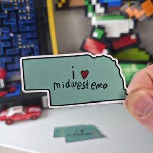 May include: A sticker in the shape of the state of Nebraska, with a light green background and a black outline. The sticker reads "i heart midwest emo" in black text with a red heart.