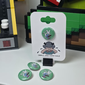 May include: A set of green buttons with a cartoon rabbit surfing on a pink surfboard. The buttons are displayed on a white card with the text "SMALL SAD YARD SALE" and a crying cartoon character.