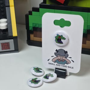 May include: White button pins with a cartoon alligator design and the text "SMALL SAD YARD SALE". The alligator is green with a red mouth and purple tongue. The pins are on a white card with a crying cartoon character.