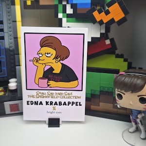 May include: A rectangular print featuring Edna Krabappel from The Simpsons, with a pink background. She is depicted with a cigarette, holding a pink mug, and wearing a black shirt that says "bright eyes". The print is part of "The Springfield Collection".