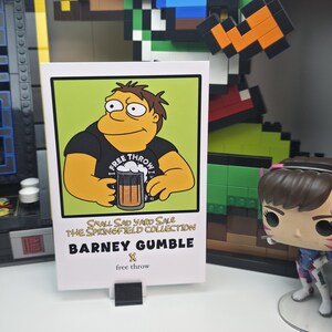 May include: A white card featuring Barney Gumble from The Simpsons, holding a beer. The card has a green background and the text "FREE THROW". Below the image, it reads "SMALL SAD YARD SALE THE SPRINGFIELD COLLECTION BARNEY GUMBLE free throw".