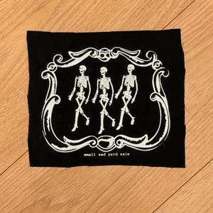 May include: Black fabric patch featuring three white skeleton figures walking in a row, framed by a decorative white border. The text "small sad yard sale" is printed in white at the bottom of the patch.
