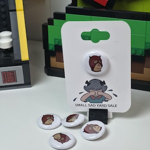 May include: A collection of round, white buttons with cartoon illustrations. One button is attached to a white display card with the text "SMALL SAD YARD SALE". The buttons feature a sad-faced character in a brown mask.
