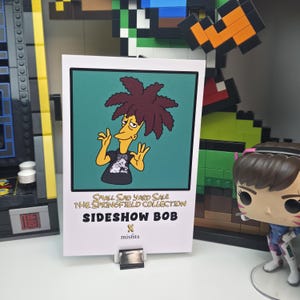 May include: A white card featuring Sideshow Bob from The Simpsons, with a green background. The card reads "SMALL SAD YARD SALE THE SPRINGFIELD COLLECTION SIDESHOW BOB x misfits". The character has red hair and is wearing a black shirt.