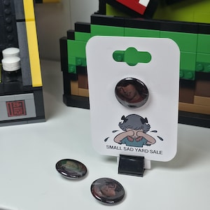 May include: A set of three round buttons with a glossy finish. One button is attached to a white card with a cartoon illustration and the text "SMALL SAD YARD SALE". The buttons feature a portrait image.