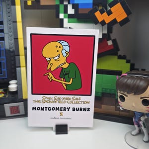May include: A white card featuring a cartoon image of Mr. Burns from The Simpsons, set against a red background. The card includes the text "Small Sad Yard Sale The Springfield Collection Montgomery Burns x Indian Summer."