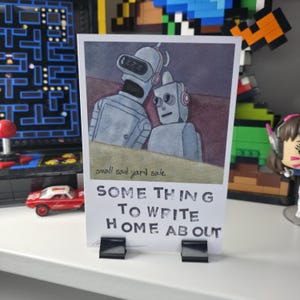 May include: A print featuring two robots with the text "SOME THING TO WRITE HOME ABOUT" and "small sad yard sale." The robots are gray and the print is held by two black clips.