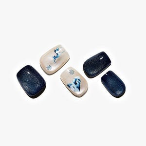 May include: Five artificial nails. Two are a dark blue with glitter, and three are a light beige with blue winter-themed designs. One nail has a blue deer, another has a snowflake, and the third has a blue heart.