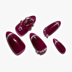 May include: A set of six artificial nails in a deep burgundy color. Some nails feature silver tips and decorative embellishments, including small silver beads and purple gems. One nail has a seashell design.