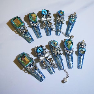 May include: A set of ten long, blue glitter press-on nails. Each nail is adorned with ornate silver embellishments and large, colorful gemstones. Some nails feature a blue eye design, and one set is connected by a silver chain.