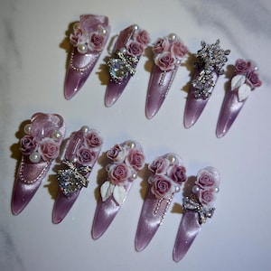 Könnte beinhalten: Ein Set aus zehn langen, mandelförmigen, lavendelfarbenen Kunstnägeln. Jeder Nagel ist mit dekorativen Elementen verziert, darunter kleine rosa Rosen, Perlen und funkelnde Strasssteine. Einige Nägel weisen eine silberne Schleife oder eine Ansammlung von Kristallen auf.