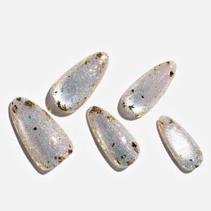 May include: Five almond-shaped artificial nails. Each nail is a light, iridescent color with gold foil accents. The nails are arranged on a white surface, casting soft shadows.