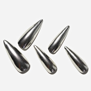 Könnte beinhalten: Sechs silberfarbene, längliche Nagelspitzen sind auf weißem Hintergrund angeordnet. Jede Spitze hat ein spitzes Ende und eine abgerundete Basis, die das Licht reflektiert. Das metallische Finish verleiht ihnen ein poliertes, modernes Aussehen. Für Nageldesign.