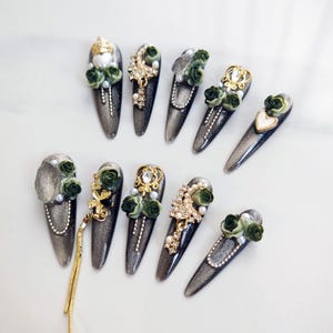 May include: A set of ten long, almond-shaped artificial nails. Each nail is painted with a shimmering gray polish and adorned with various embellishments, including green roses, pearls, and gold or silver charms. Some nails feature a dangling gold chain.