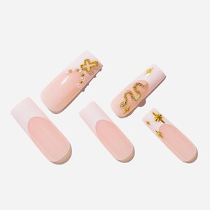 May include: A set of five artificial nails. The nails have a light pink base with a white tip. Two nails feature gold embellishments: a cross and a snake. The other three nails have a simple French tip design.