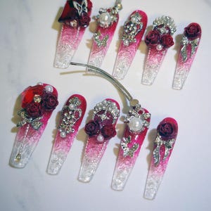 May include: A set of ten long, artificial nails with a gradient pink and clear design. Each nail is adorned with decorative elements like red roses, pearls, and silver rhinestones. Some nails feature a butterfly or bow accents.