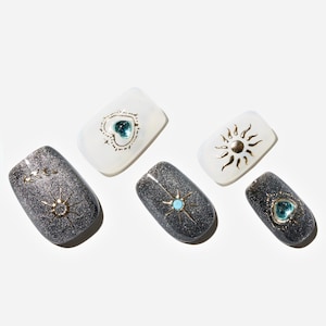 May include: A collection of artificial nails in various designs. Some nails are a glittery dark gray, adorned with gold sunbursts and turquoise or gold accents. Others are white with gold sun and heart designs.