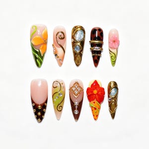 Peut inclure: Un ensemble de dix faux ongles en forme d'amande présentant des motifs variés, incluant des fleurs en relief, des volutes dorées, des pois marron, des strass et des textures métalliques complexes dans des tons orange, marron, vert et or.