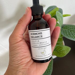 Puede incluir: Una botella de vidrio marrón con un gotero negro y una etiqueta blanca que dice "HORMONE BALANCE SALVIA OFFICINALIS HERBAL SUPPLEMENT 2 FL OZ (60ml) Alchemy Care Med".