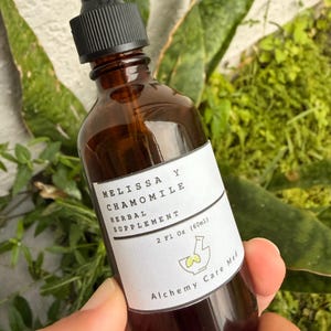 May include: An amber glass dropper bottle containing a herbal supplement labelled Melissa y Chamomile, 2 Fl Oz (60ml), with a black dropper cap and a white label featuring a mortar and pestle icon and the text Alchemy Care Med.