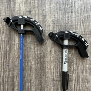 May include: Two black 3D-printed angle finders with degree markings, each holding a writing utensil. One holds a blue pencil, the other a black Sharpie marker. The tools are on a wooden surface.