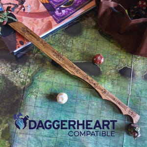 May include: A wooden Daggerheart compatible sword with engraved geometric designs. The sword is lying on a green and blue map with dice and a brown bag. The text "Daggerheart Compatible" is visible.