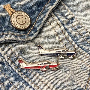 May include: Two silver aeroplane pins on a blue denim background. The top pin is blue and white with the text "N4KRLW". The bottom pin is red, white, and blue with the text "N4KRLW".