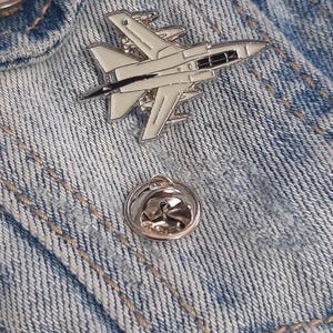 May include: A white enamel pin of a fighter jet on a blue denim jacket.